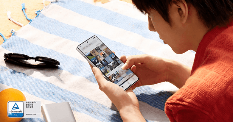 A person using a OnePlus 13 smartphone while sitting on a striped beach towel, with sunglasses and an orange nearby.