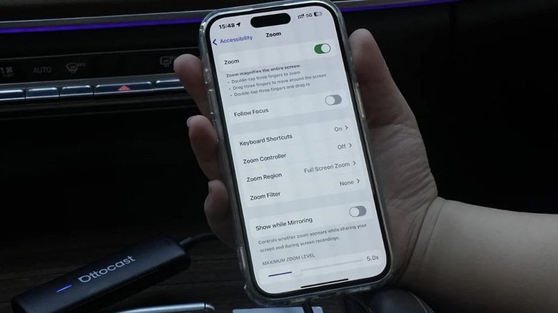 A hand holding a phone displaying Zoom settings under Accessibility, with an Ottocast device nearby. A hand holding a phone displaying Zoom settings under Accessibility, with an Ottocast device nearby.