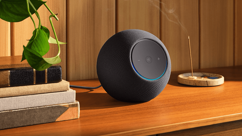 Ein schwarzer Amazon Echo Studio Lautsprecher auf einer Holzoberfläche mit Büchern und einer Pflanze im Hintergrund.