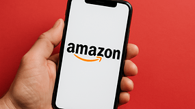Eine Hand hält ein Smartphone, das das Amazon-Logo auf einem weißen Bildschirm vor rotem Hintergrund zeigt.