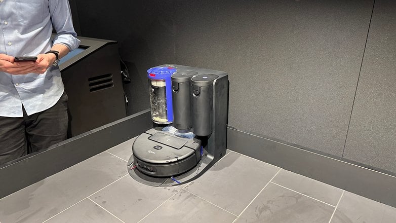 A Dyson Spot+Scrub Ai robotic cleaner on a tiled floor, with a person holding a phone nearby.