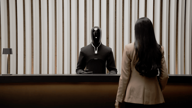 Ein humanoider Roboter an einem Empfangstresen, der mit einer Frau im Anzug interagiert.
