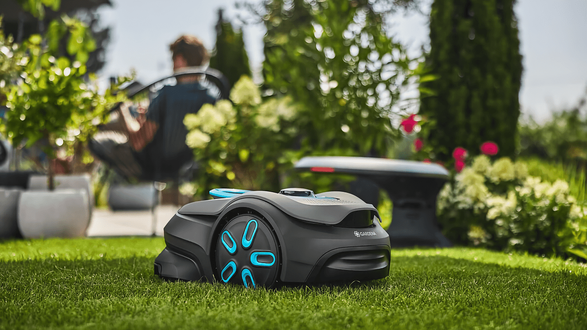 Un robot de tonte Gardena Smart Sileno Sense sur de l'herbe verte avec une personne se relaxant en arrière-plan.