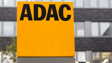 Ein großes gelbes Schild mit dem Text 'ADAC' vor einem verschwommenen Hintergrund von Fensterfassaden.