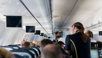 Interior of an airplane cabin with rows of seats and overhead screens, a flight attendant serving passengers.