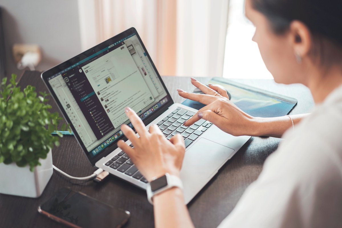 Una mujer tecleando en una laptop con una pequeña planta y un teléfono inteligente en la mesa.