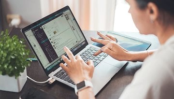 Eine Frau tippt auf einem Laptop mit einer kleinen Pflanze und einem Smartphone auf dem Tisch.