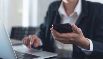 Eine Frau sitzt am Laptop und schaut auf ihr Handy