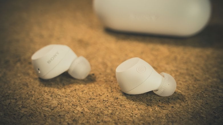 Fones de ouvido Sony brancos em uma superfície de cortiça com um estojo de carregamento ao fundo. Fones de ouvido Sony brancos em uma superfície de cortiça com um estojo de carregamento ao fundo.