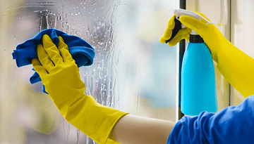 Eine Person putzt ein Fenster mit einem blauen Tuch und einer Sprühflasche.