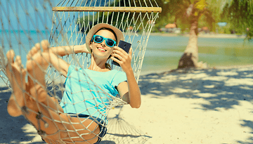 Eine Frau, die in einer Hängematte am Strand entspannt, ein Smartphone benutzt und Sonnenbrillen trägt.