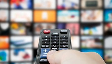 Eine Hand hält eine Fernbedienung vor einer Wand von Fernsehern, die verschiedene Inhalte anzeigen.