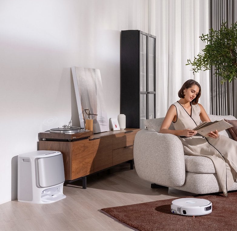 Eine Frau liest eine Zeitschrift auf einem Sofa, während ein Staubsauger-Roboter auf dem Boden steht.