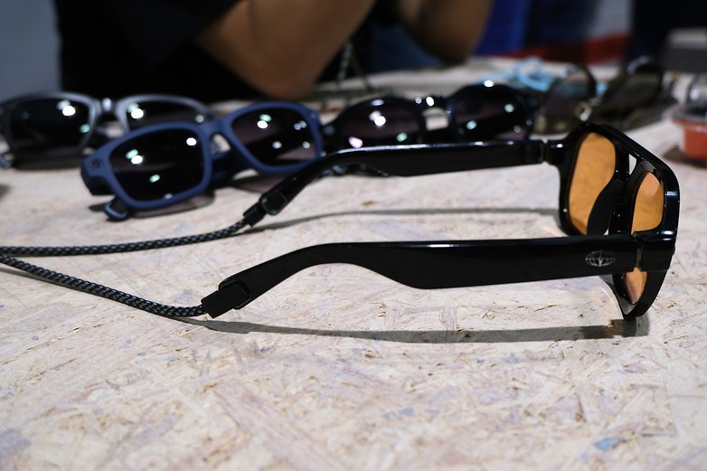 A pair of black sunglasses with orange lenses, laying on a wooden surface among other sunglasses.