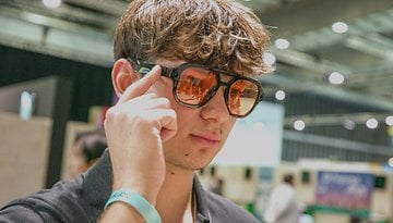 A young man wearing stylish sunglasses, touching his temple, in an indoor event space.
