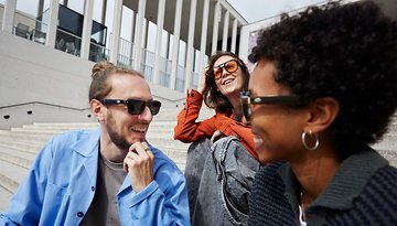 Drei Freunde lachen und tragen Sonnenbrillen bei einer Veranstaltung auf Stufen.